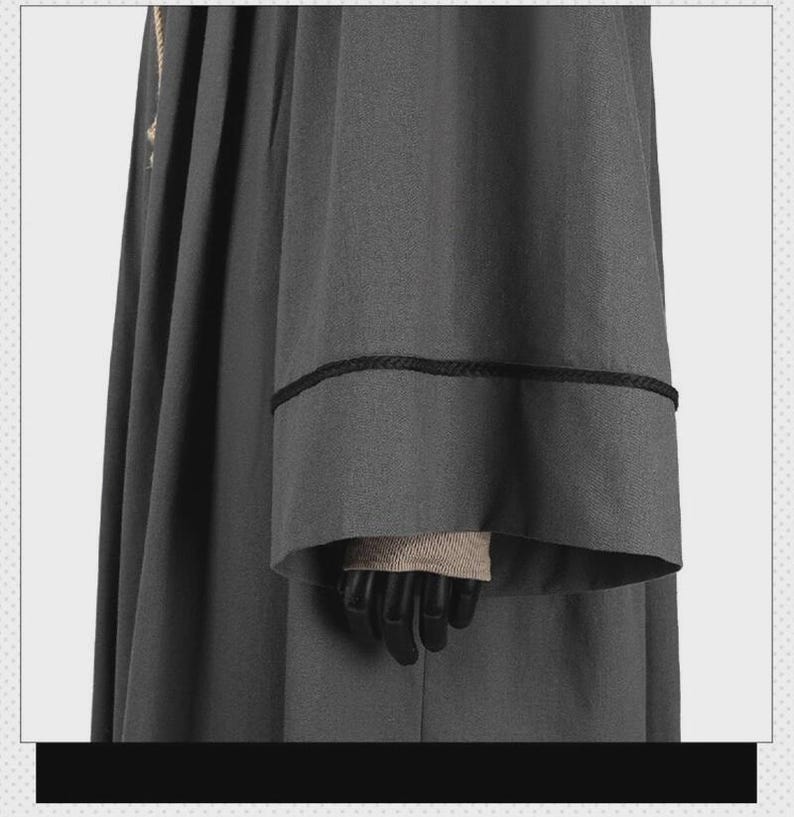 May include: A dark gray robe featuring long, wide sleeves with a black braided trim detail around the cuffs. The garment has a structured, pleated design with a vertical black braided trim running along the side of the main body fabric.