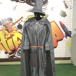 May include: A grey wizard costume featuring a long-sleeved robe with a matching cape and a tall pointed hat. The outfit includes a brown belt with a silver buckle and a dark grey pouch attached at the waist. A gold-colored decorative clasp secures the cape.