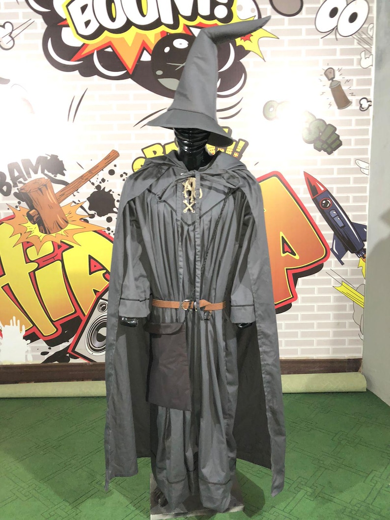 May include: A grey wizard costume featuring a long-sleeved robe with a matching cape and a tall pointed hat. The outfit includes a brown belt with a silver buckle and a dark grey pouch attached at the waist. A gold-colored decorative clasp secures the cape.