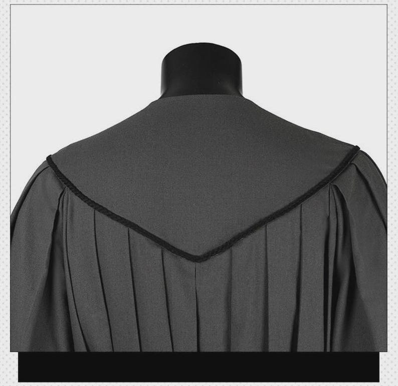 May include: A charcoal gray academic gown featuring a V-shaped yoke across the back and shoulders. The garment is constructed with vertical pleats extending from the yoke down the back, finished with a thin black corded trim along the edges.