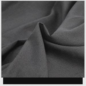 May include: A dark charcoal gray fabric with a visible twill weave texture. The material is draped to create soft folds and shadows, highlighting the matte finish and the diagonal pattern of the weave across the surface of the textile.