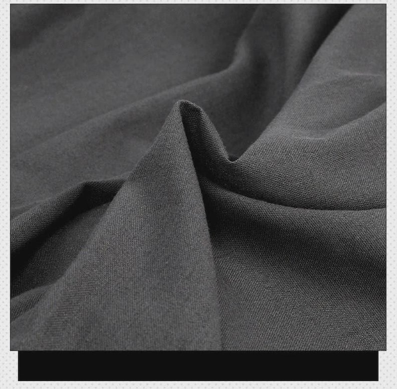 May include: A dark charcoal gray fabric with a visible twill weave texture. The material is draped to create soft folds and shadows, highlighting the matte finish and the diagonal pattern of the weave across the surface of the textile.