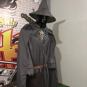 May include: A grey wizard costume featuring a tall pointed hat, a long flowing cape, and a tunic with a lace-up neckline. The outfit includes a brown belt with a buckle and a dark brown pouch attached at the waist, displayed on a black mannequin.