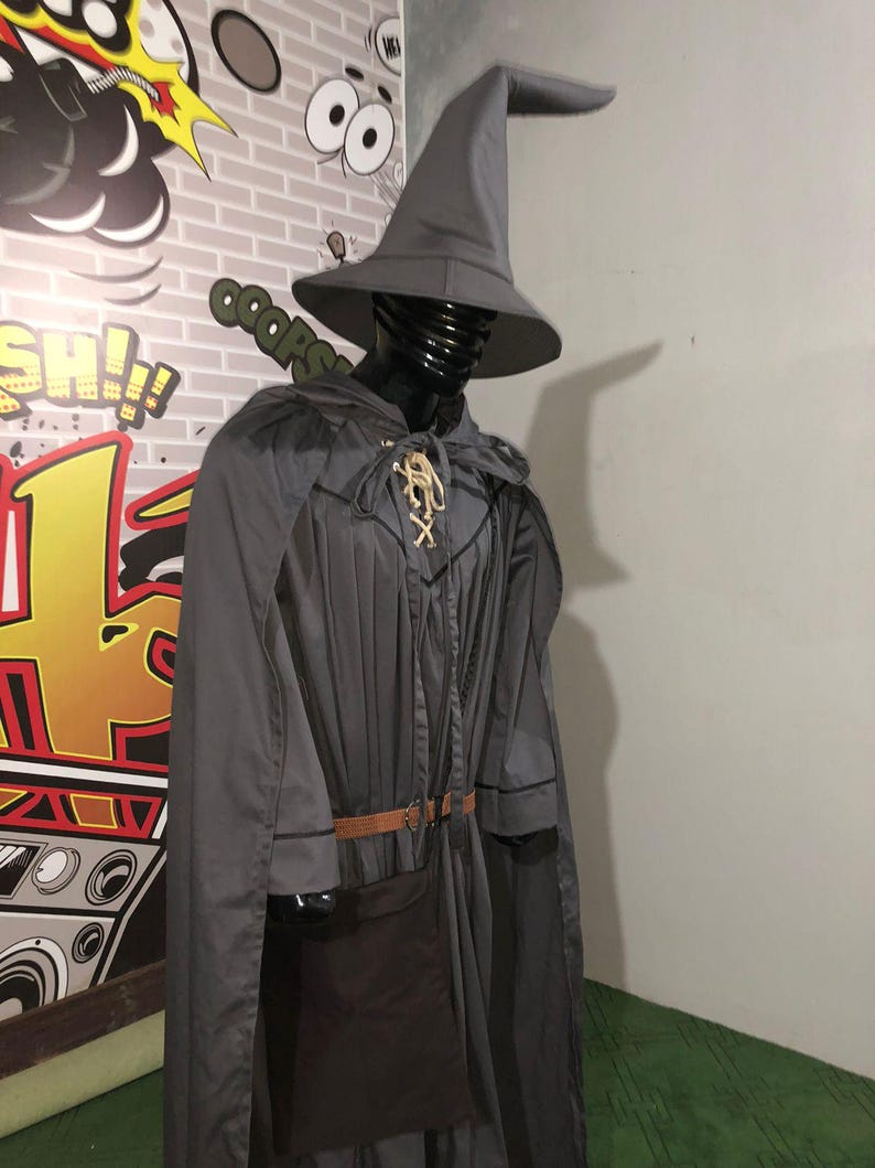 May include: A grey wizard costume featuring a tall pointed hat, a long flowing cape, and a tunic with a lace-up neckline. The outfit includes a brown belt with a buckle and a dark brown pouch attached at the waist, displayed on a black mannequin.