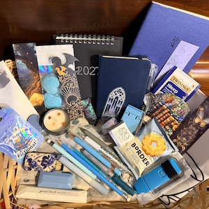 May include: A gift basket containing a blue notebook with a gothic window design, a 2023 calendar, various bookmarks with fantasy illustrations, a small candle, pens, a box of matches, Twinings Sleep tea, Border shortbread, and stationery items.