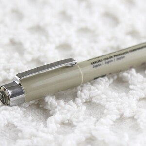 Op de afbeelding: Een beige Sakura Color Products Copic marker met een zilveren dop en het zwarte nummer "01" op de dop. De marker ligt op een wit gehaakt oppervlak.