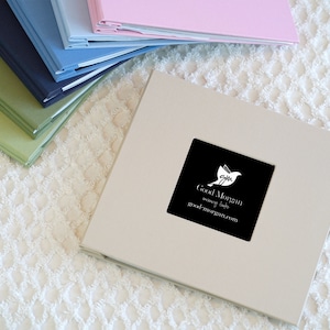May include: A beige photo album with a black square sticker on the front. The sticker features a white bird with a black outline and the text "Good Morgan" in white lettering. The text "memory book" and "goodmorgan.com" are also in white lettering.