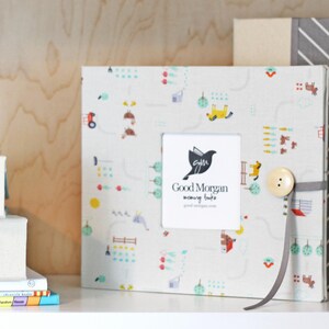 May include: A white photo album with a colorful pattern of cars, houses, and trees. The album has a small frame with the text "Good Morgan mommy hugs" and a button closure. The album is sitting on a shelf with other items.