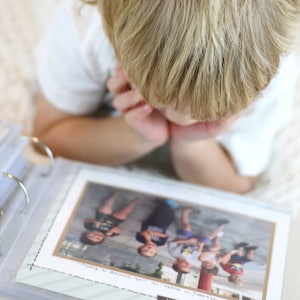 Puede incluir: Un ni&ntilde;o peque&ntilde;o mirando un &aacute;lbum de fotos lleno de fotos de un grupo de ni&ntilde;os. El &aacute;lbum de fotos est&aacute; abierto en una p&aacute;gina con una foto de cinco ni&ntilde;os de pie en fila.