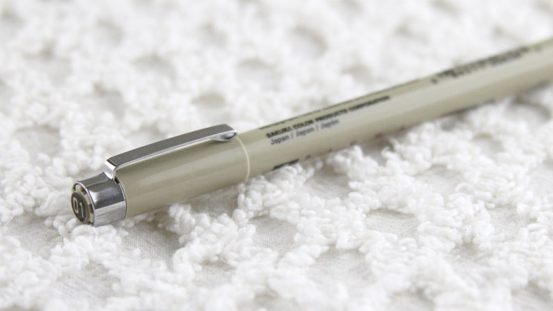 Op de afbeelding: Een beige Sakura Color Products Copic marker met een zilveren dop en het zwarte nummer "01" op de dop. De marker ligt op een wit gehaakt oppervlak.