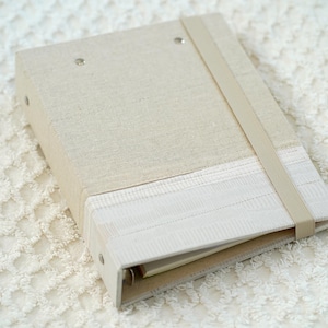 May include: A beige linen binder with a white lace trim and a beige ribbon closure.