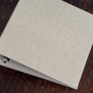 May include: A beige binder with a textured cover and a metal ring mechanism. The binder is closed and lying flat on a brown surface.