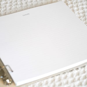 May include: Open white guest book with lined pages and the word "GUESTS" printed at the top. The book is held together with a silver three-ring binder. The book is open on a white textured surface.
