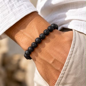 Könnte beinhalten: Ein Armband bestehend aus runden, porösen schwarzen Lavasteinperlen, die in einer Reihe aufgereiht sind. Die Perlen weisen eine texturierte, löchrige Oberfläche auf, die für Vulkangestein charakteristisch ist und ein einheitliches, dunkles Erscheinungsbild erzeugt.