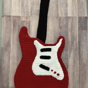 Può includere: Un oggetto da appendere a forma di chitarra realizzato a mano all'uncinetto, con corpo rosso, lungo manico nero e battipenna bianco. Il battipenna presenta tre accenti rettangolari neri orizzontali e tre piccoli dettagli circolari rossi.