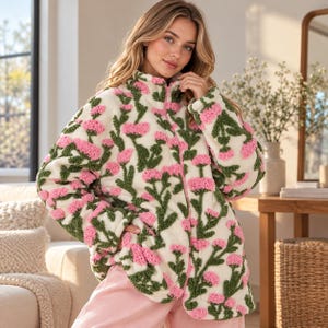 May include: A cream-colored fleece jacket featuring a high collar, a full-length front zipper, and a textured floral pattern with pink flower heads and green stems. The jacket has long sleeves and a relaxed fit, crafted from a soft, fuzzy material.