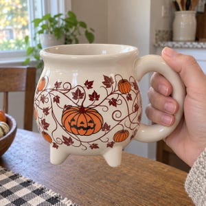 Może przedstawiać: Kremowy ceramiczny kubek w kształcie kociołka z trzema małymi nóżkami i dużym uchem. Zewnętrzna strona ozdobiona jest powtarzającym się wzorem pomarańczowych dyń i lampionów, otoczonych ciemnobrązowymi wijącymi się pnączami oraz liśćmi klonu.