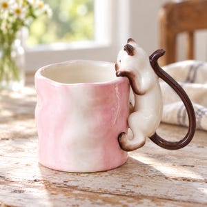 Peut inclure: Une tasse en céramique avec une finition marbrée rose clair et blanche ondulée. L'anse est en forme de chat siamois au pelage crème avec des oreilles, des pattes et une queue brun foncé, semblant grimper sur le bord et le long de la tasse.