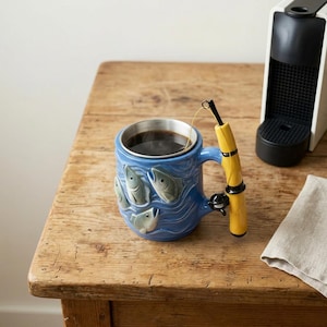 Puede incluir: Una taza de cerámica azul con un relieve de peces nadando en el exterior. El asa tiene forma de caña de pescar amarilla con un carrete negro. Un hilo de pescar fino se extiende desde la caña hacia el líquido oscuro dentro de la taza.