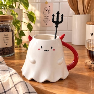 May include: A white ceramic mug shaped like a ghost with small red horns and a red handle. The mug features a cute face with black eyes, a wavy mouth, and pink cheeks. A black trident-shaped spoon rests inside the mug.