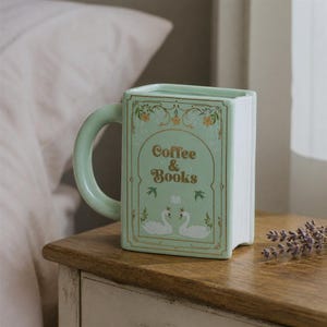 May include: A light green ceramic mug shaped like a rectangular book. The front features gold text reading Coffee & Books, gold floral accents, and two white swans wearing crowns. The side of the mug has vertical ridges resembling book pages.