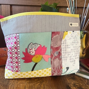 May include: A fabric pouch featuring a patchwork design with a central light blue panel displaying a pink and green flower appliqué. The top section is grey with thin white stripes and a yellow trim. A small label reads Stitch Nests.