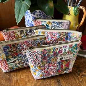 May include: A collection of four zipped pouches made from patchwork fabric featuring various floral patterns in red, blue, yellow, and green. Each pouch has a metal zip and a small white fabric label with the text Stitch Nests.