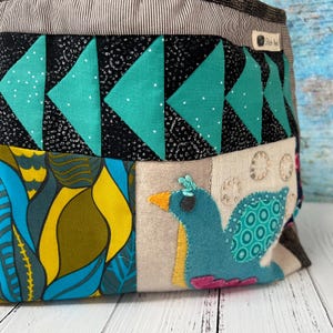 May include: A fabric storage basket featuring a patchwork design with a top band of black and teal triangles, a section of yellow and blue abstract leaf patterns, and a beige panel with a teal bird appliqué and the label Shish Nest.