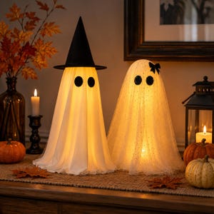 May include: Two illuminated ghost decorations. The left ghost wears a tall black witch hat and has two black circular eyes. The right ghost is draped in sheer white fabric with subtle glitter and features a small black bow and two black eyes.
