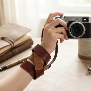 May include: A dark brown leather camera wrist strap wrapped multiple times around a wrist. The strap features a smooth finish on the outer band and a textured, grainy finish on the inner section, connected to a silver and black digital camera.