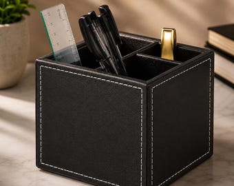 May include: A square black desk organizer with white contrast stitching, featuring three interior compartments. It holds two black pens, a clear plastic ruler marked in inches, and a gold-toned metal object. The organizer has a textured finish.