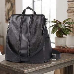 May include: A black backpack with a smooth central panel flanked by two vertical braided accents. The bag features a top handle, adjustable shoulder straps with silver-toned metal hardware, and side pockets on both the left and right sides.