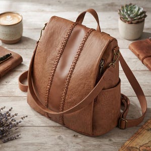 May include: A medium brown backpack featuring a smooth central vertical panel flanked by two braided leather-like strips. The bag includes a top handle, adjustable shoulder straps with antique brass-toned hardware, and side pockets with zippers.