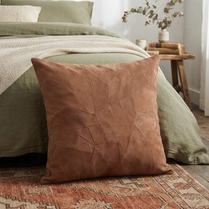 May include: A square throw pillow in a solid terracotta brown color. The pillow features a patchwork design composed of multiple suede-like fabric pieces stitched together in an irregular geometric pattern across the entire front surface.
