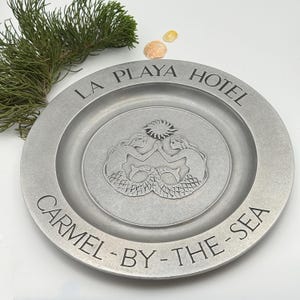 May include: A round, silver-toned metal plate featuring an embossed design of two mermaids with a sun above them. The text LA PLAYA HOTEL is engraved at the top, and CARMEL-BY-THE-SEA is engraved along the bottom rim of the plate.