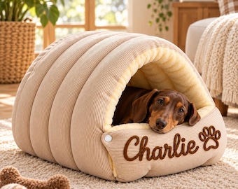 May include: A beige corduroy pet bed with a tunnel-like shape and a cream-colored interior lining. The front features the embroidered name Charlie in brown script next to a brown paw print icon. A brown dachshund rests inside the bed.