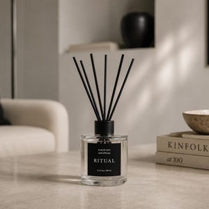 Op de afbeelding: Een glazen rietdiffuser met een zwart etiket met de tekst RITUAL, CLEAN DAY, reed diffuser, 3.4 fl oz | 100 ml. De fles heeft een zwarte dop en bevat verschillende dunne zwarte rietjes die vanuit de opening in het midden omhoog steken.