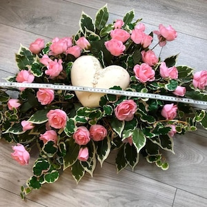 Könnte beinhalten: Ein cremefarbenes herzförmiges Ornament, eingebettet zwischen rosa Rosen und panaschierten grün-weißen Blättern. Ein weißes Maßband liegt über dem Gesteck und zeigt eine Breite von etwa 60 cm von einem Ende zum anderen.