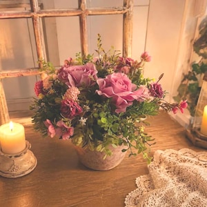 Può includere: Una composizione floreale in un vaso rotondo e testurizzato con rose rosa, fiori più piccoli e fogliame verde. La composizione è esposta accanto a una candela accesa in un supporto bianco anticato e un pezzo di tessuto in pizzo.
