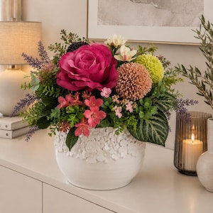 Op de afbeelding: Een bloemstuk met een grote magentakleurige roos, een perzikkleurige pomponbloem, een limoengroene bolbloem en kleine roze bloesems met groen blad. De bloemen zitten in een witte keramische vaas met een gestructureerd bloemenpatroon.