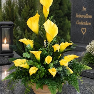 Könnte beinhalten: Ein Blumenarrangement aus leuchtend gelben Calla-Lilien und grünem Blattwerk in einem runden Topf. Es steht auf einer dunklen Steinfläche neben einer schwarzen Laterne und einem Grabstein mit der Inschrift In liebevollem Gedenken und einem Herz.