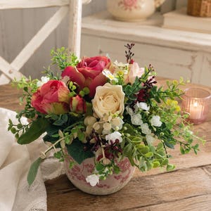 Könnte beinhalten: Ein Blumenarrangement in einem runden, rosafarbenen Keramiktopf mit Blumenmuster. Der Strauß besteht aus dunkelrosa und cremefarbenen Rosen, kleinen weißen Blüten, dunkelroten Beeren und verschiedenen grünen Blattstielen in dichter Anordnung.
