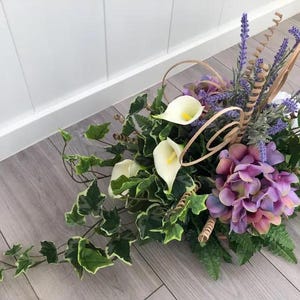 Pode incluir: Um arranjo floral com lírios de calla brancos, flores de hortênsia roxas e ramos de alfazema. O conjunto inclui folhas de hera verdes com bordas claras, folhas de feto e detalhes decorativos de madeira enrolada integrados.