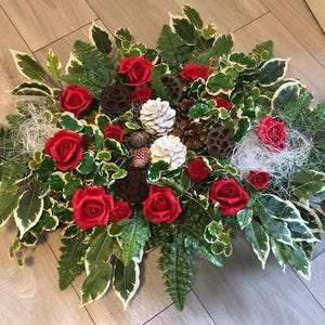 Pode incluir: Um arranjo floral com rosas vermelhas vibrantes, pinhas com pontas brancas, vagens de sementes de lótus castanho-escuras e folhas variegadas em verde e branco. A composição inclui fibras brancas finas e folhas de feto verdes.