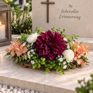 Könnte beinhalten: Ein Blumengesteck mit einer großen dunkelburgunderfarbenen Dahlie, weißen Ranunkeln, pfirsichfarbenen Eukalyptusblättern und kleinen weißen Füllblumen auf einer Moosbasis, platziert vor einem Grabstein mit Kreuz und der Inschrift In liebevoller Erinnerung.