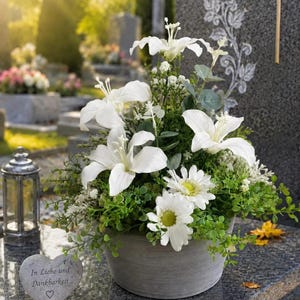 Könnte beinhalten: Ein Blumenarrangement in einem grauen Topf mit weißen Lilien, weißen Gänseblümchen und grünem Blattwerk. Ein kleiner herzförmiger Stein liegt daneben, auf dessen Vorderseite der Text In Liebe und Dankbarkeit und ein Herzsymbol eingraviert sind.