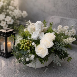 Könnte beinhalten: Eine weiße Keramik-Engelsfigur mit betenden Händen, eingebettet in ein Blumenarrangement aus weißen Rosen, grünen Beeren und verschiedenem Grün in einer weißen Schale, begleitet von einer schwarzen Laterne mit einer brennenden Kerze.
