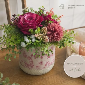 Op de afbeelding: Een bloemstuk in een cilindervormige keramische pot met een roze bloemenpatroon. Het boeket bevat een grote magentakleurige roos, een oudroze chrysant, kleine witte bloemetjes en groen blad. De tekst op de afbeelding luidt "Für jedes Zuhause geeignet" en "HANDARBEIT mit Liebe".