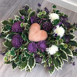 Peut inclure: Un arrangement floral décoratif composé d'une pierre centrale lisse en forme de cœur couleur pêche, entourée de fleurs en copeaux de bois violet foncé, de fleurs blanches type chrysanthème et de feuilles vertes et blanches panachées.