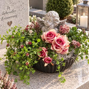 Op de afbeelding: Een bloemstuk in een zwarte, geribbelde schaal met roze rozen, kleine witte en rode bloemen en groen blad. Een grijs stenen engeltje staat tussen de bloemen op een grijze stenen ondergrond.